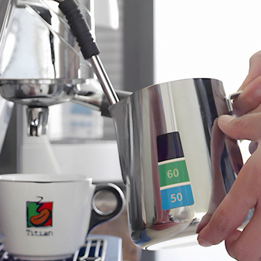 Person using a digital milk frothing pitcher with a Barista coffee machine in the background.